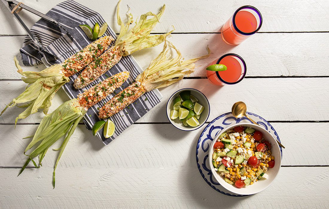 elote and corn salad on blue dishes rest on a white wooden table with drinks and limes