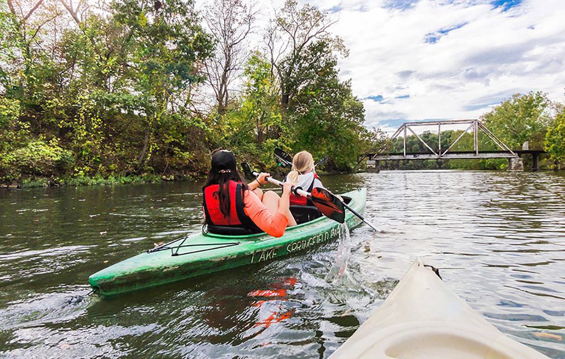 Two girls paddle their kayak down the river towards a bridge on James River in Springfield, MO