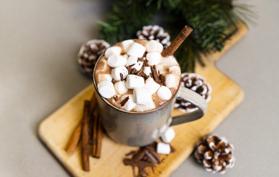 A mug of hot cocoa topped with marshmallows, chocolate pieces and a cinnamon stick rests on a wooden platter decorated with garland, cinnamon sticks and pinecones.