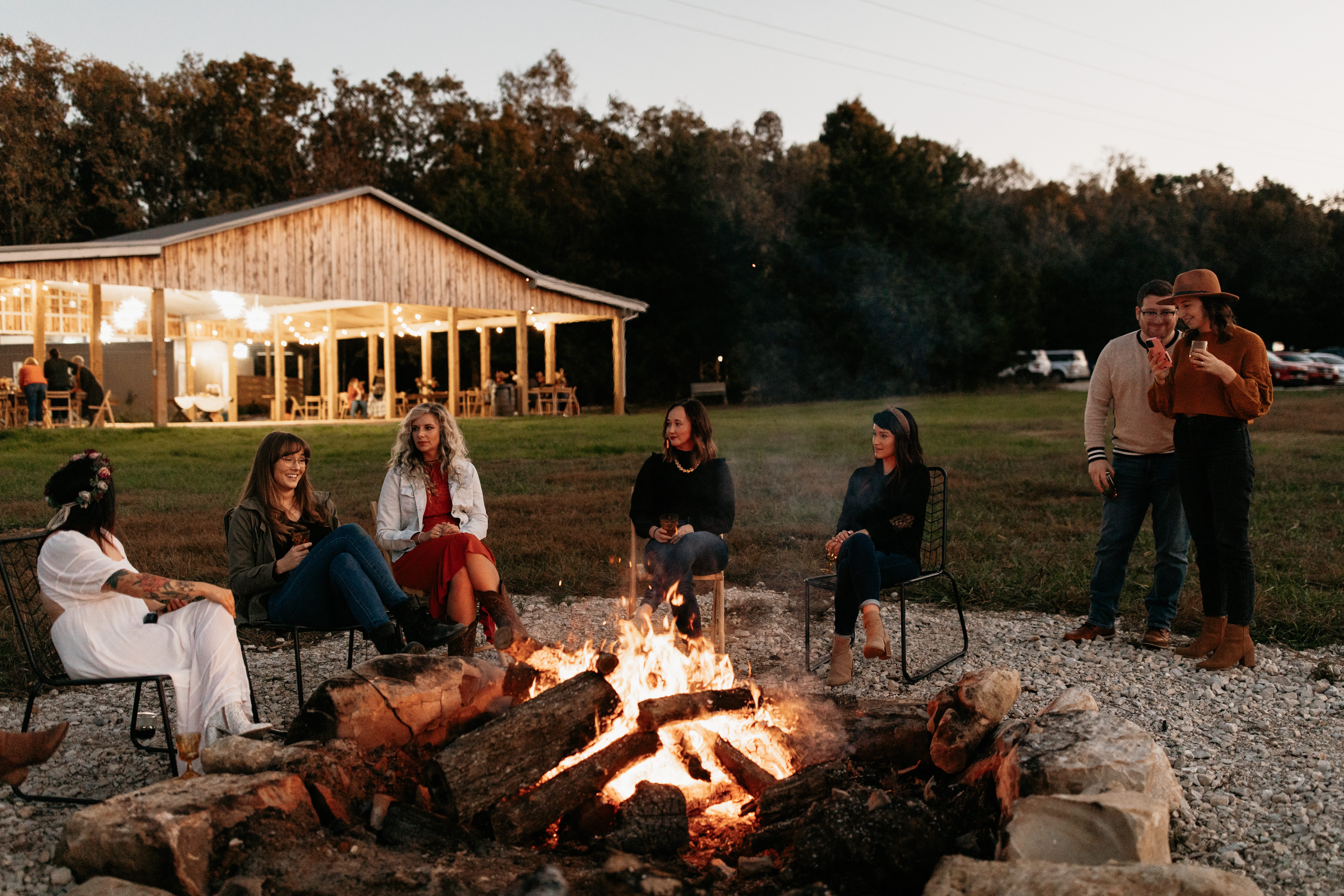 A group of guests talks and laughs around a fire pit at sunset, with a warmly lit barn in the far background.