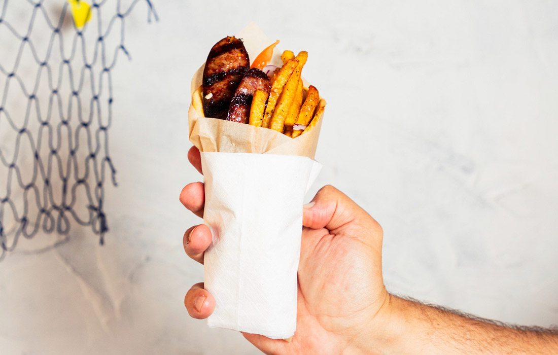 A hand holds up a pita wrap against a white backdrop. Fries, charred sausage and tomato stick out of the the wrap.