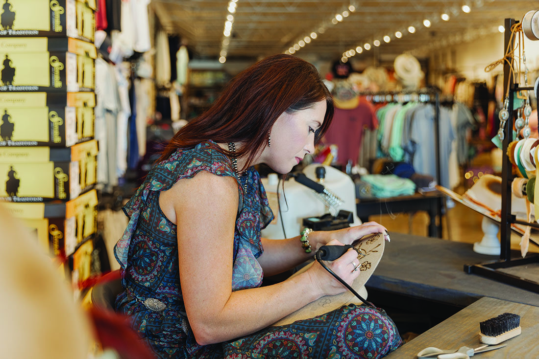 A woman burns designs into a Western-style hat with a specialized tool.