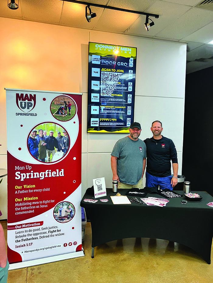 Two men smile behind a table, set up with brochures and large sign about Man Up and Go.