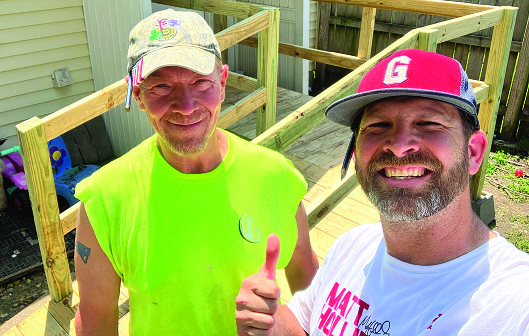Two men in work clothes and ball caps smile at the camera. One flashes a thumbs-up.