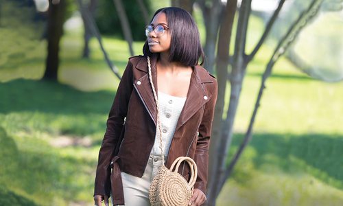 girl stands in brown jacket and tan purse in a park