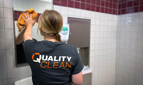 A Quality Clean employee wipes down a mirror in a commercial restroom.
