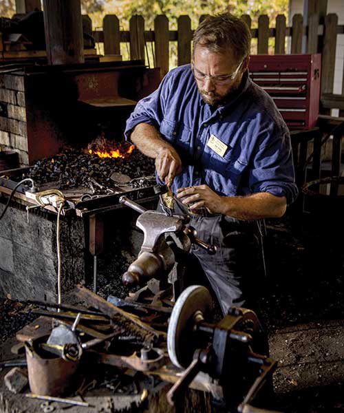 A man, Sam Hibbs, work with tools at the mill. An assortment of metal pieces and tools are organized around him.