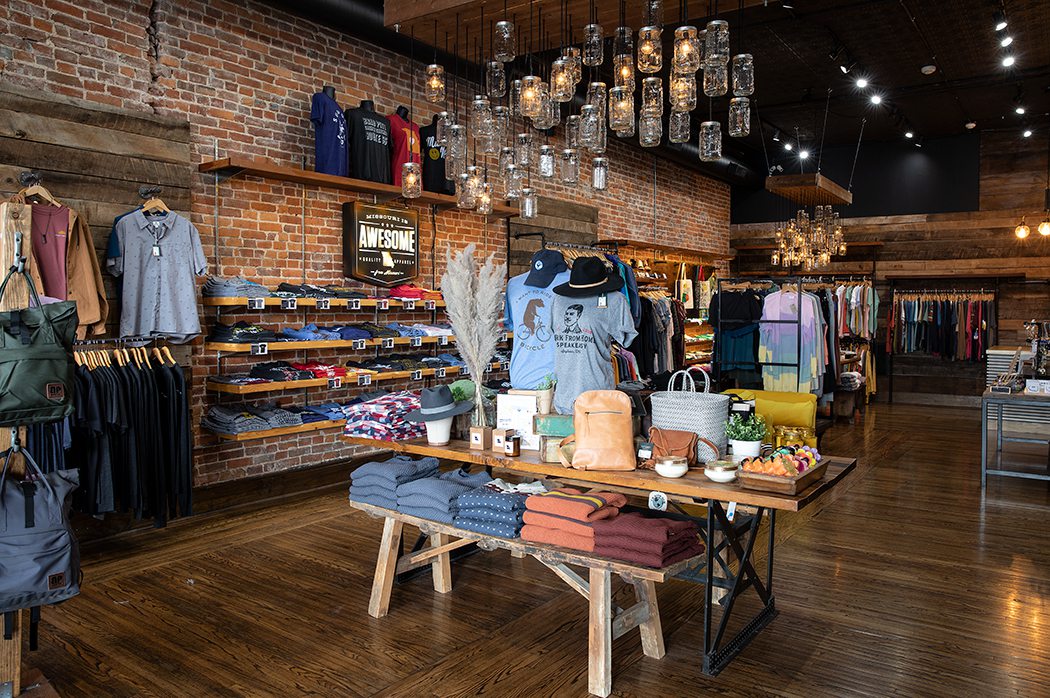 Clothing racks and table of folded clothing in 5 Pound Apparels new location downtown with wooden floors, brick walls, and unique light fixtures and decor