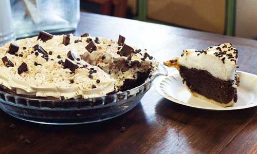 A slice of chocolate pie covered in whipped cream sits on a plate next to the rest of the pie