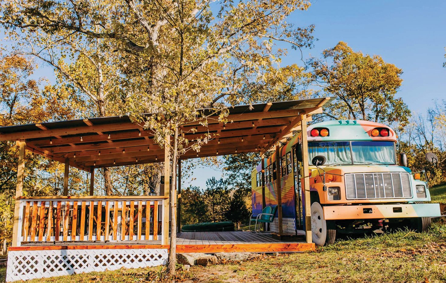 Exterior photo of a colorful school bus converted into a camper parked beside a large wooden deck