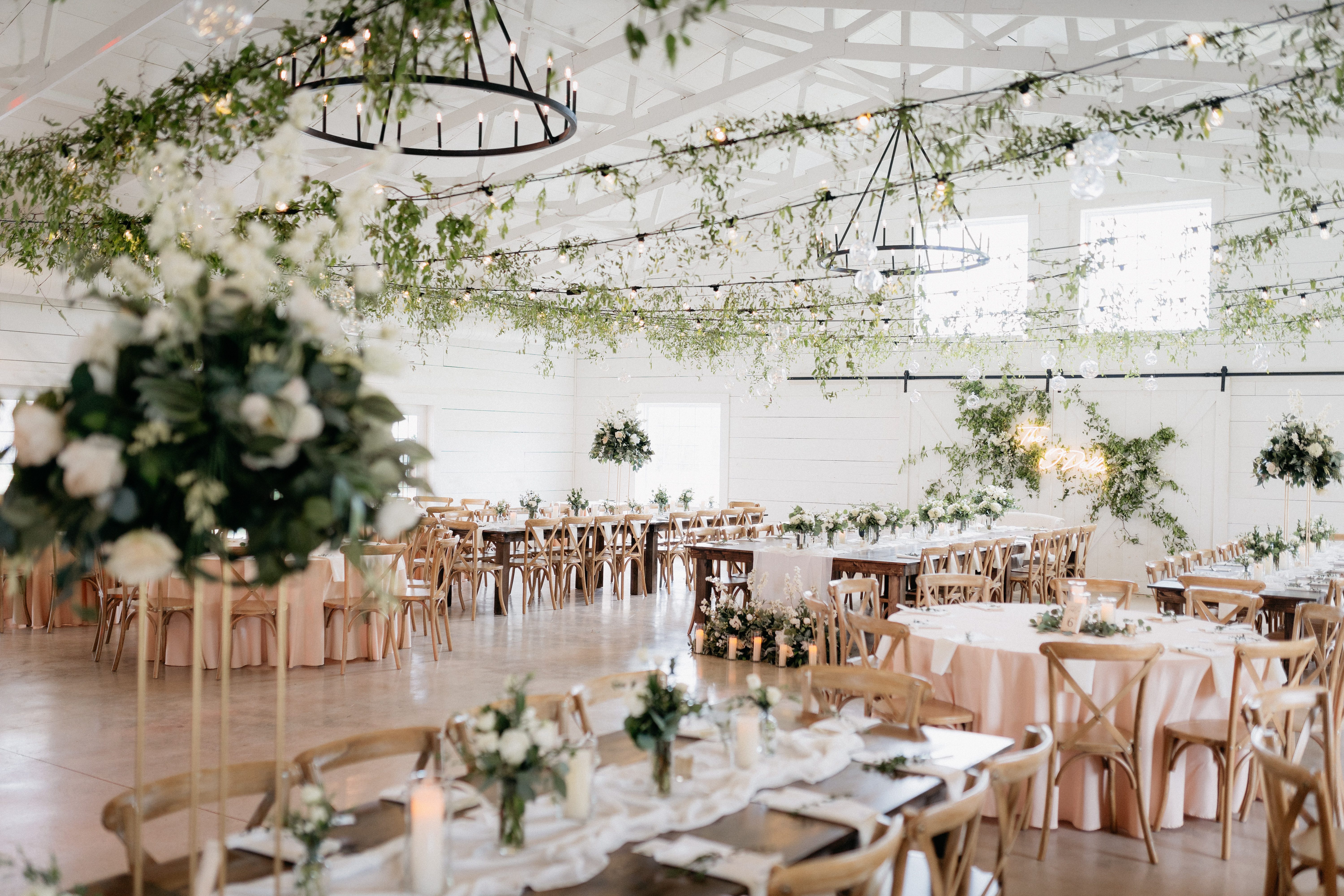 White lights and greenery are strung across rafters inside a bright, airy barn. Satin tablecloths, votives and floral arrangements top each long reception table.