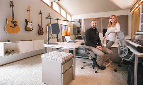Phil sits and Chastin stands in their clean office room; guitars line the walls