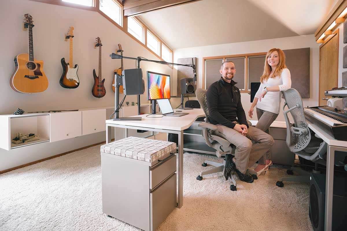 Phil sits and Chastin stands in their clean office room; guitars line the walls