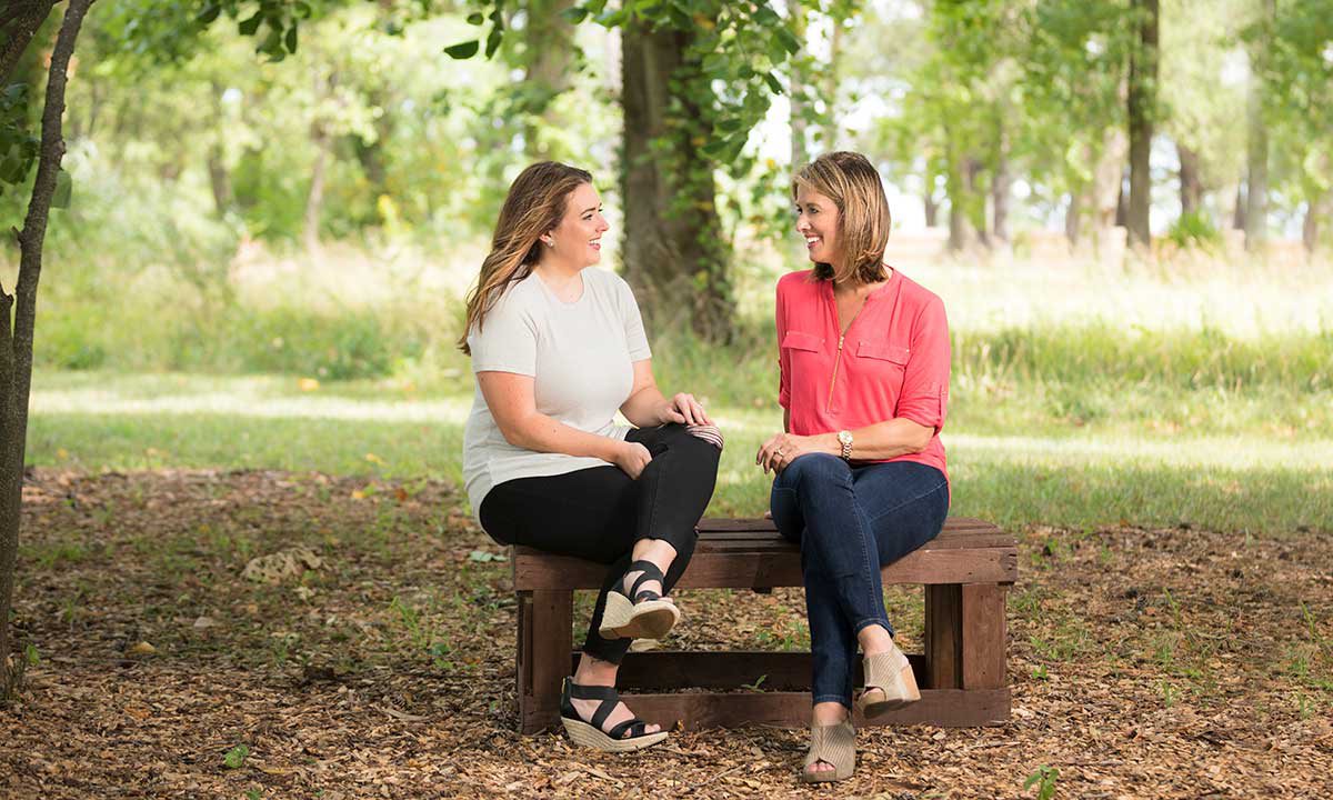 Jody and Julie sit on a bench speaking in a park