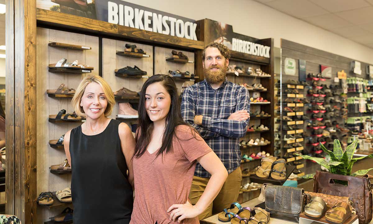 Two women and a man pose and smile in front of rows of shoes