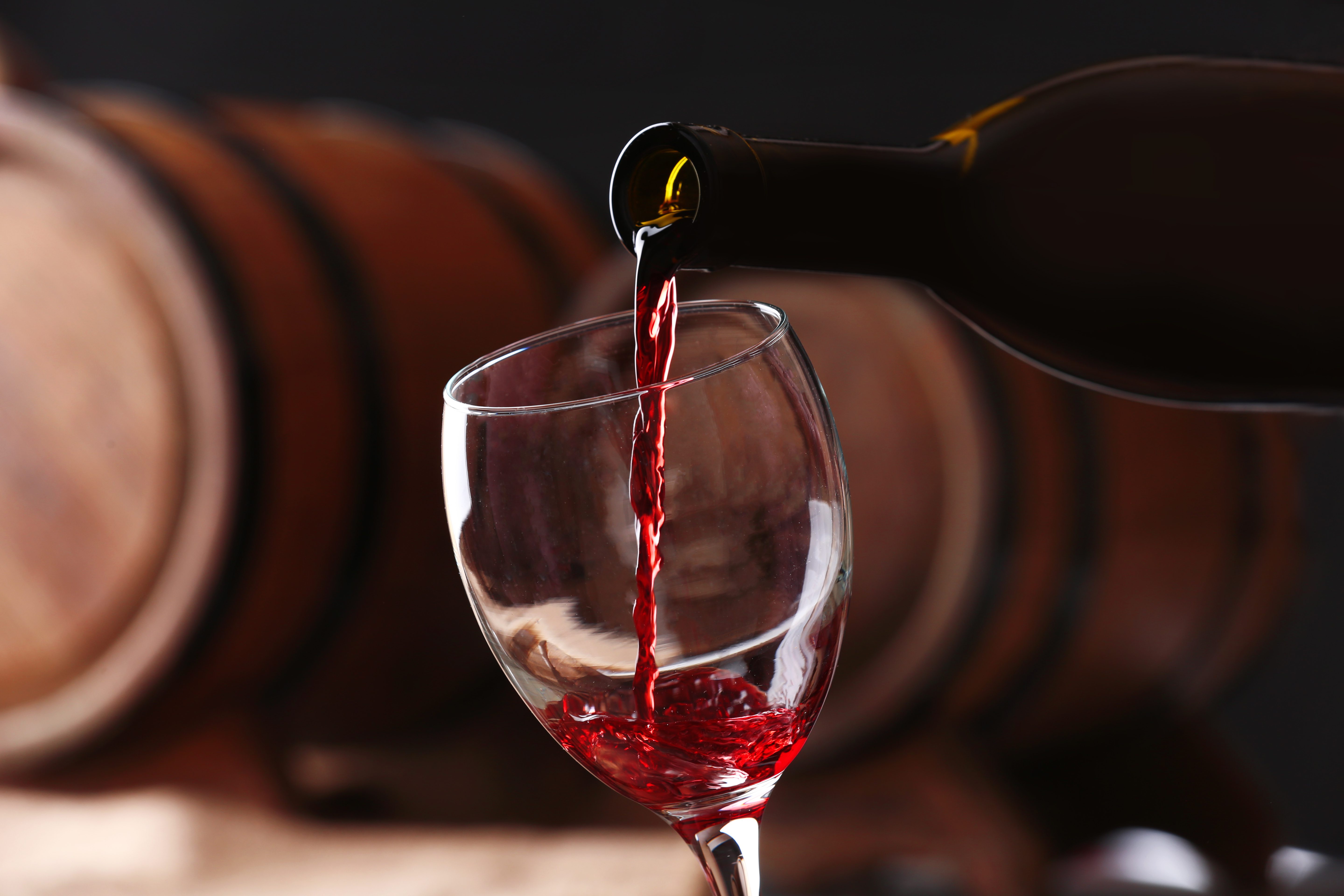 Red wine is poured into a glass against a background of stacked barrels.