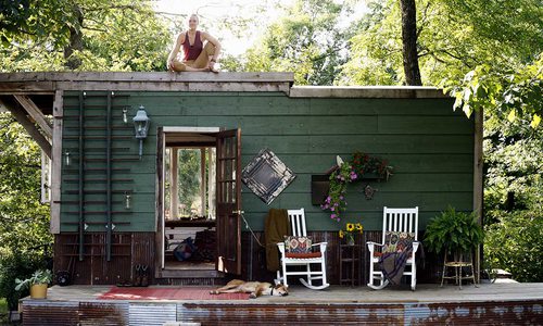 Jayme sits on top of the tiny home, her dog lounges on the base